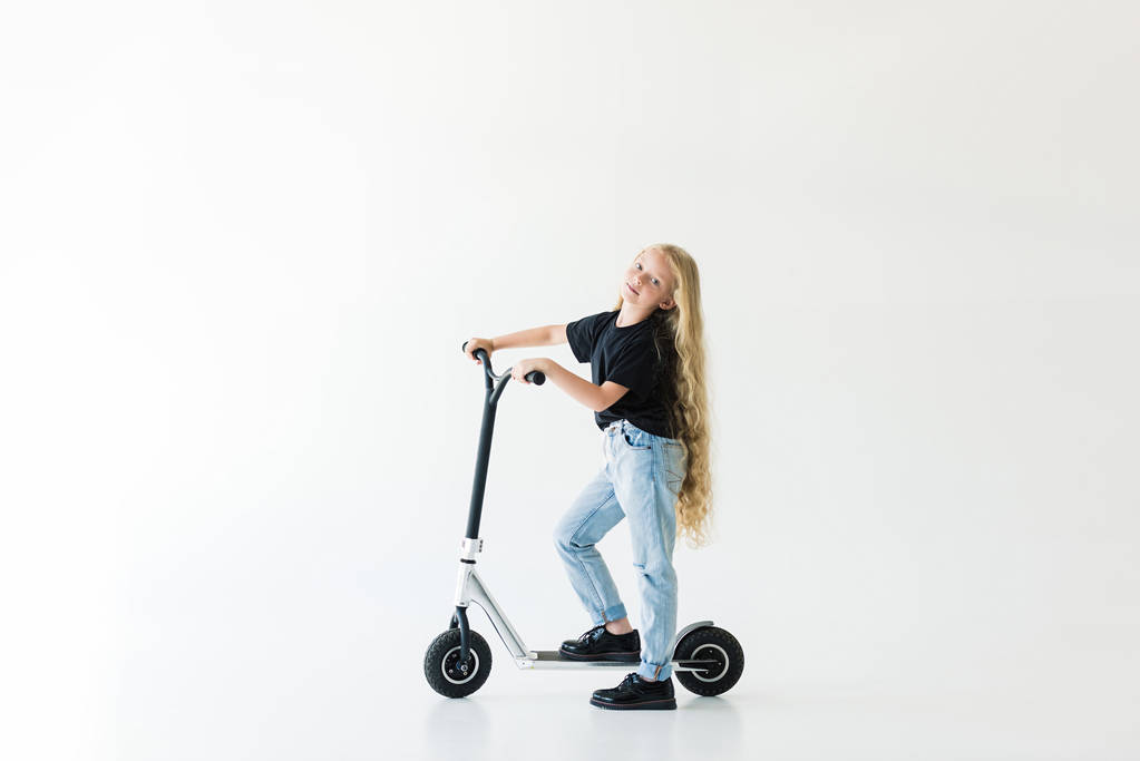 Вид ребенка в полный рост с длинными кудрявыми волосами, который едет на скутере и смотрит на камеру, изолированную на белом фоне
  - Фото, изображение