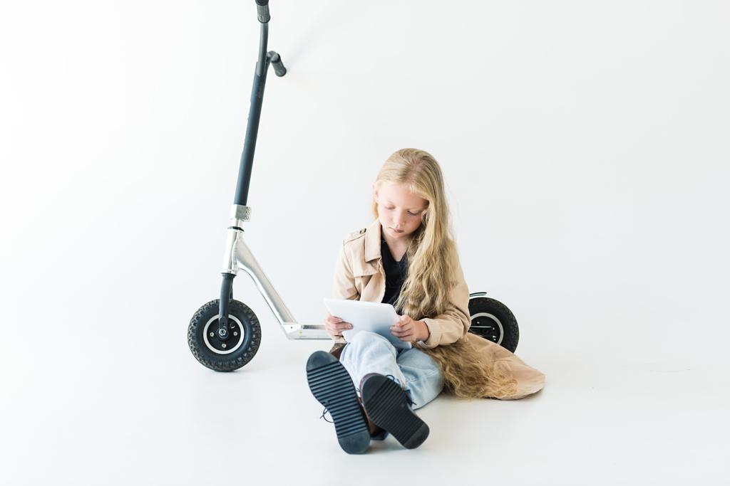 magnifique enfant avec de longs cheveux bouclés en utilisant une tablette numérique tout en étant assis près de scooter isolé sur blanc
  - Photo, image