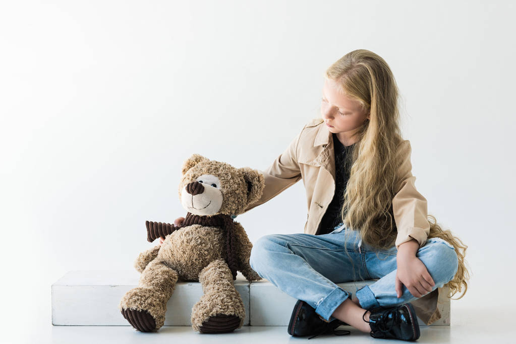 full length view of cute stylish child sitting and looking at teddy bear on white  - Photo, Image