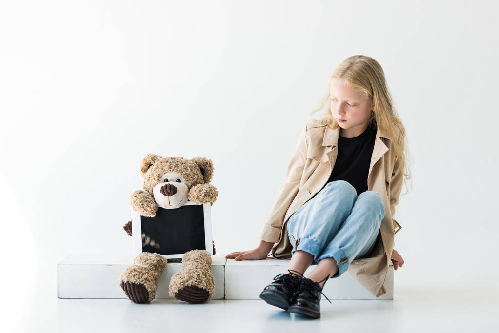schönes Kind mit langen lockigen Haaren schaut Teddybär mit digitalem Tablet auf weißem Hintergrund an - Foto, Bild
