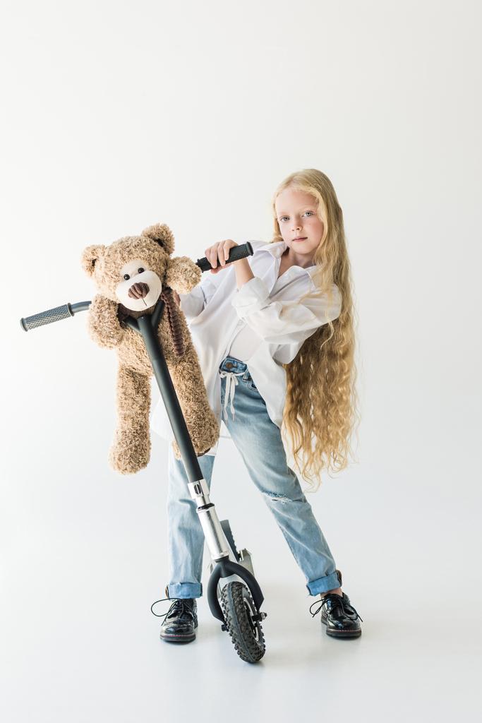 roztomilé dítě s dlouhými kudrnatými vlasy s skútr a medvídka a zadíval se na kameru na bílém  - Fotografie, Obrázek