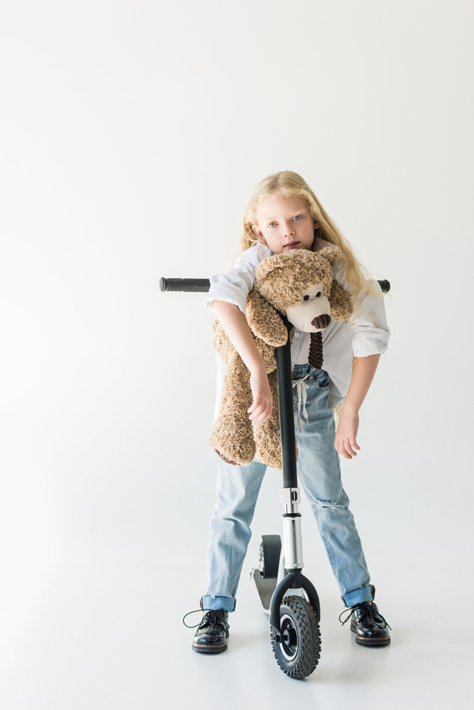 αξιολάτρευτο παιδί ακουμπά στο σκούτερ με αρκουδάκι και κοιτάζοντας την κάμερα στο λευκό - Φωτογραφία, εικόνα