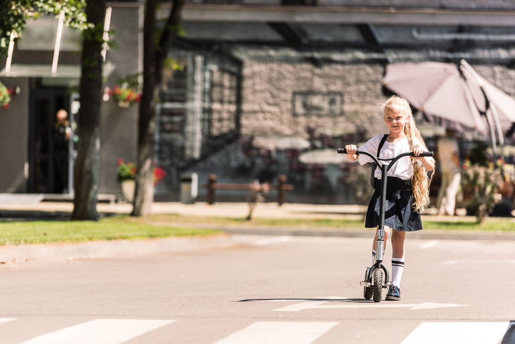 beautiful little schoolgirl with long curly hair riding scooter at sunny day - Photo, Image