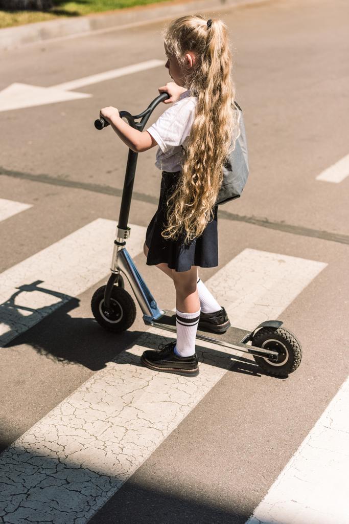 pohled z vysokého úhlu rozkošné malé školačky s dlouhé kudrnaté vlasy jezdecké skútr na ulici  - Fotografie, Obrázek