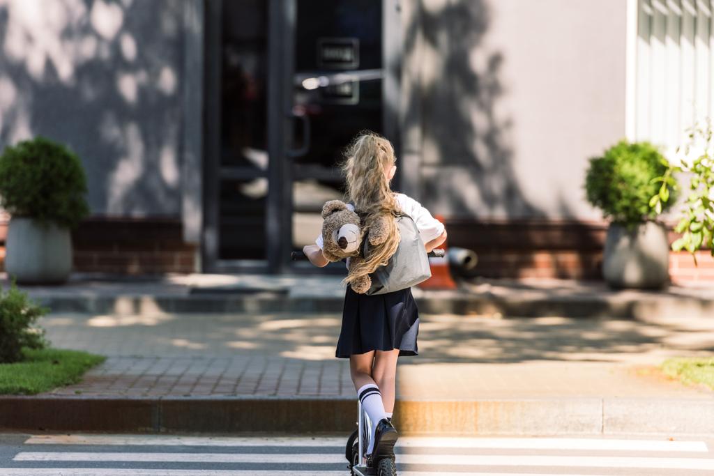 visão traseira de pequeno estudante bonito com mochila e ursinho de pelúcia montando scooter na rua
    - Foto, Imagem