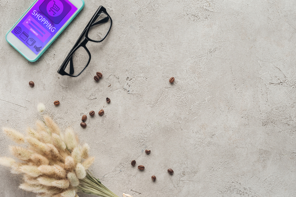 vista superior del teléfono inteligente con aplicación de compras en pantalla con gafas, granos de café derramados y ramo de lagurus ovatus en la superficie de hormigón
 - Foto, imagen