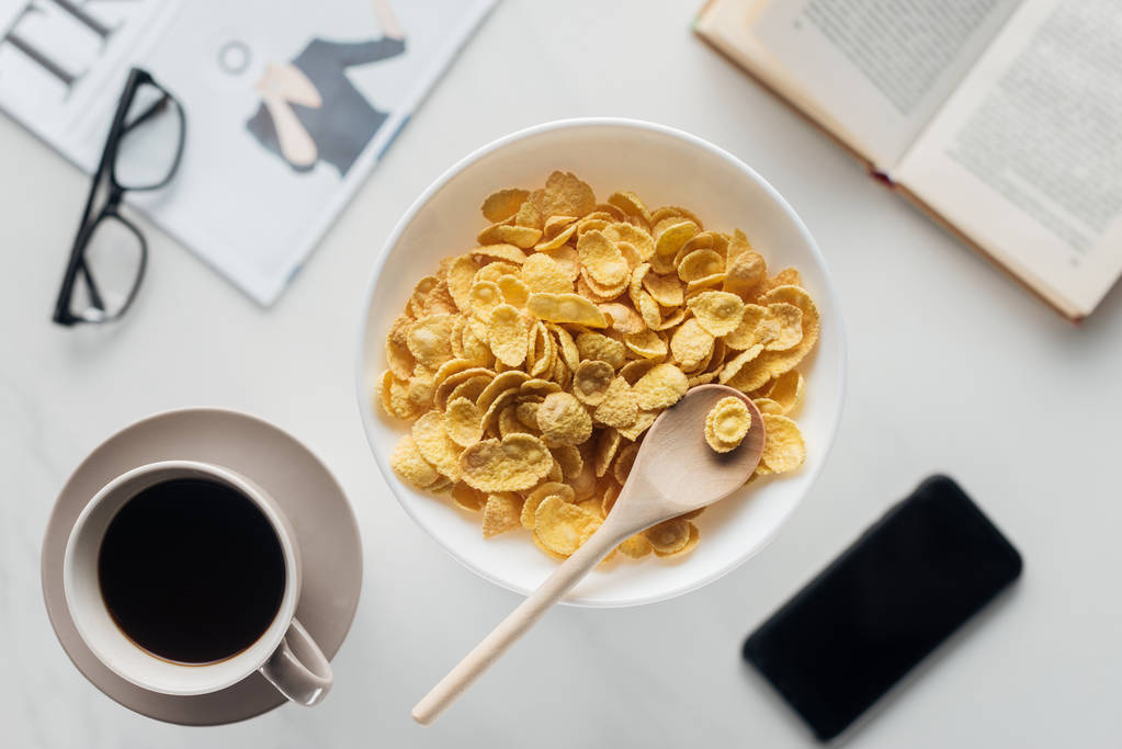 pohled shora mísy Kukuřičné vločky snídaně s šálkem kávy a croissantu na bílém povrchu s novinami, smartphone a knihy - Fotografie, Obrázek