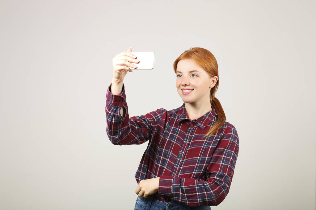 Портрет грайливої рудої жінки в фланельній картатій сорочці, яка приймає селфі з мобільним телефоном, ізольований сірий фон. Студентка тримає смартфон, фотографує. Копіювання простору, крупним планом
. - Фото, зображення