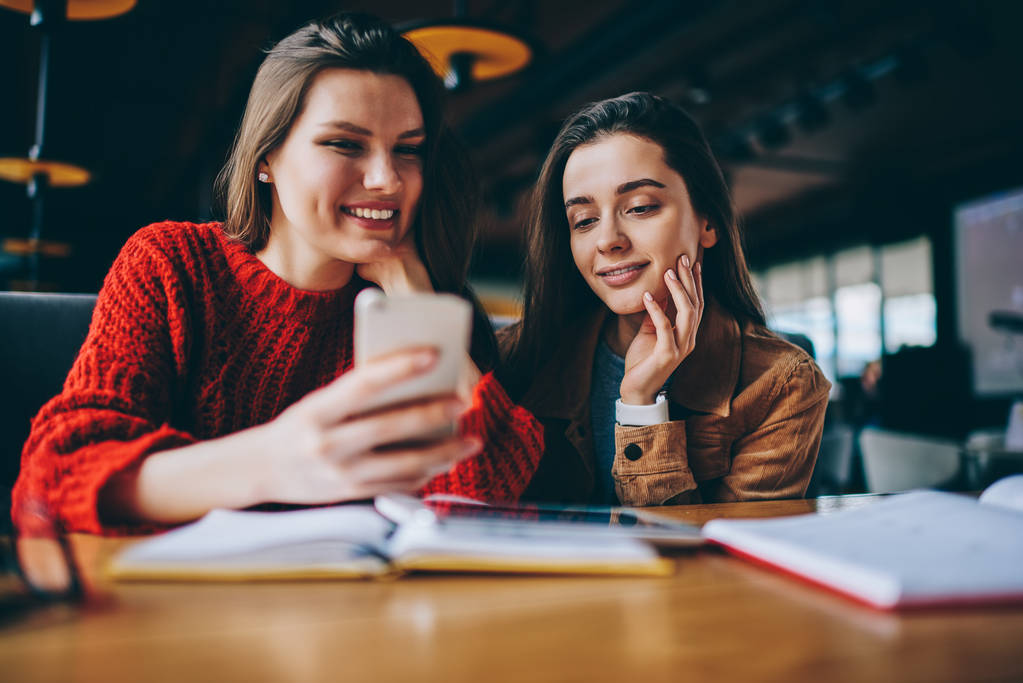 Θετική φίλες ψάχνουν πληροφορίες για εκπαίδευση χρησιμοποιώντας smartphone συσκευή και Wi-Fi σύνδεση σε καφετέρια, επιτυχόντες φοιτητές απολαμβάνοντας σπάσει μελετώντας βλέποντας αστείο βίντεο από το τηλέφωνο - Φωτογραφία, εικόνα
