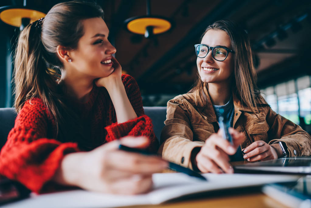 Positive Hipster-Mädchen, die die Live-Kommunikation genießen, einander zuschauen und Blickkontakt beim Lernen in Innenräumen aufnehmen, lächelnde Frauen, die gemeinsam in der Universitätsbibliothek nach Hausaufgaben forschen - Foto, Bild