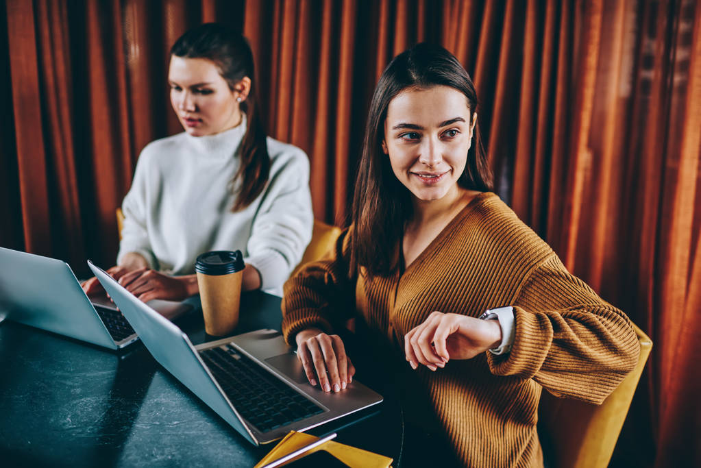 Две успешные женщины-фрилансеры, работающие дистанционно на ноутбуках, сидящих за столом в кафе, позитивная хипстерша, проверяющая время и сообщение на электронные часы, проводящие время с другом
 - Фото, изображение