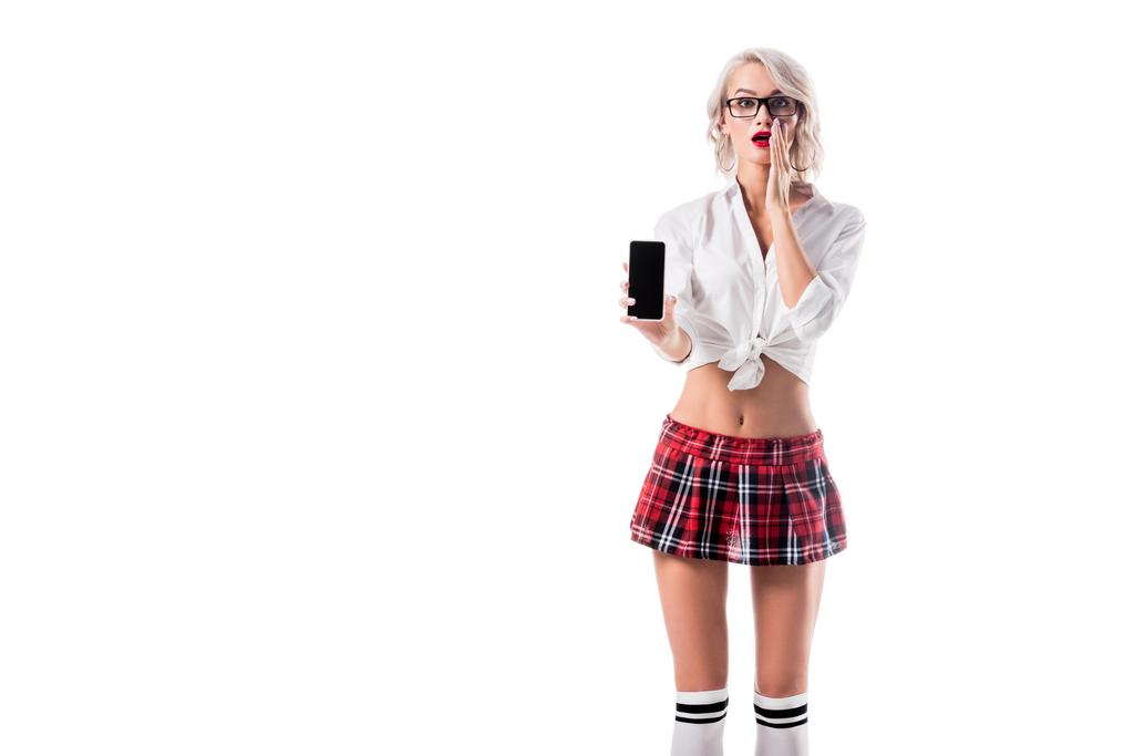 shocked woman in seductive schoolgirl clothing showing smartphone with blank screen isolated on white - Photo, Image