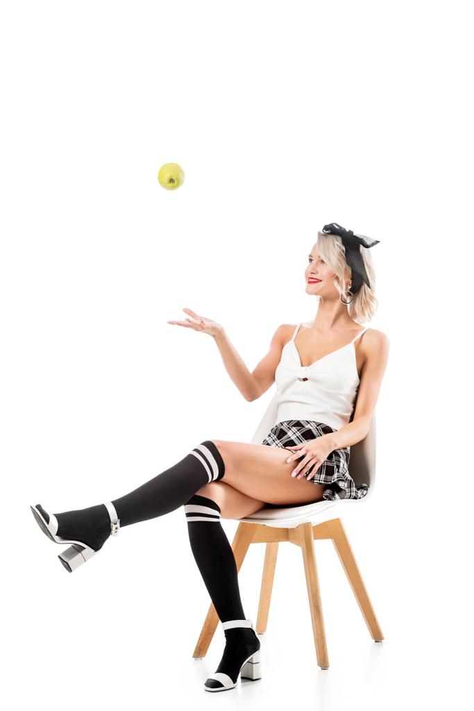 jovem sexy mulher no curto xadrez saia e joelho meias jogando maçã enquanto sentado na cadeira isolada no branco
 - Foto, Imagem
