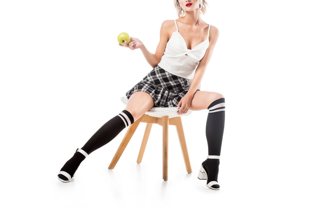 Schnappschuss einer sexy Frau in kurzem karierten Rock und Kniestrümpfen mit Apfel in der Hand, die auf einem Stuhl sitzt, der isoliert auf weiß ist - Foto, Bild