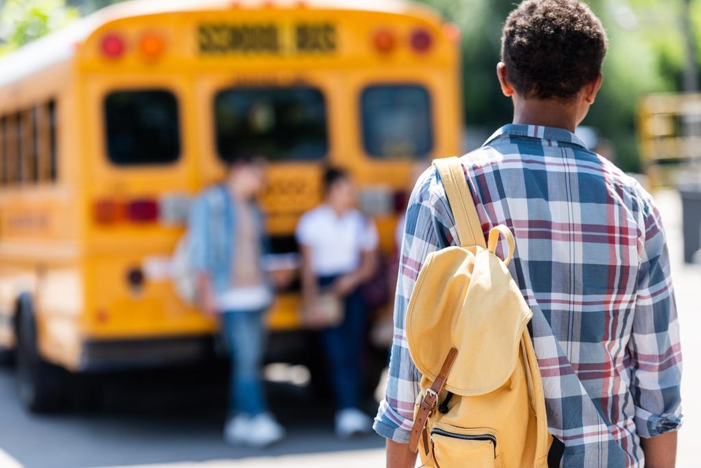 πίσω όψη του Αφρικανός Αμερικανός μαθητής περπάτημα στους συμμαθητές που ακουμπά στο σχολικό λεωφορείο - Φωτογραφία, εικόνα