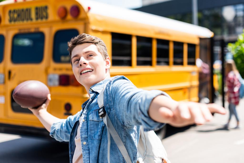 feliz adolescente escolar lanzando americano pelota de fútbol en frente de autobús escolar
 - Foto, Imagen