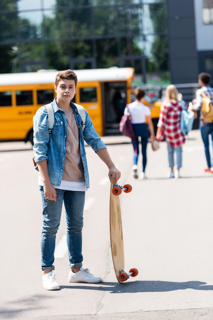 feliz adolescente escolar con monopatín de pie en frente de autobús escolar y grupo de compañeros de clase caminando borrosa en el fondo
 - Foto, Imagen