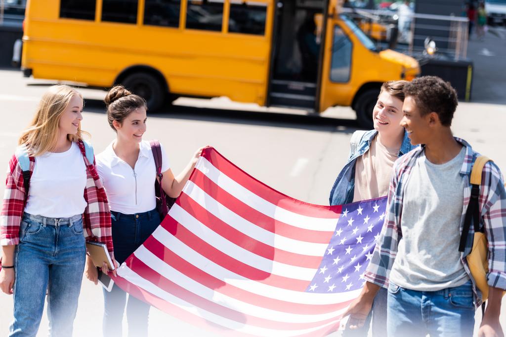 grupo de estudiosos adolescentes multiétnicos americanos con bandera de EE.UU. frente al autobús escolar
 - Foto, Imagen