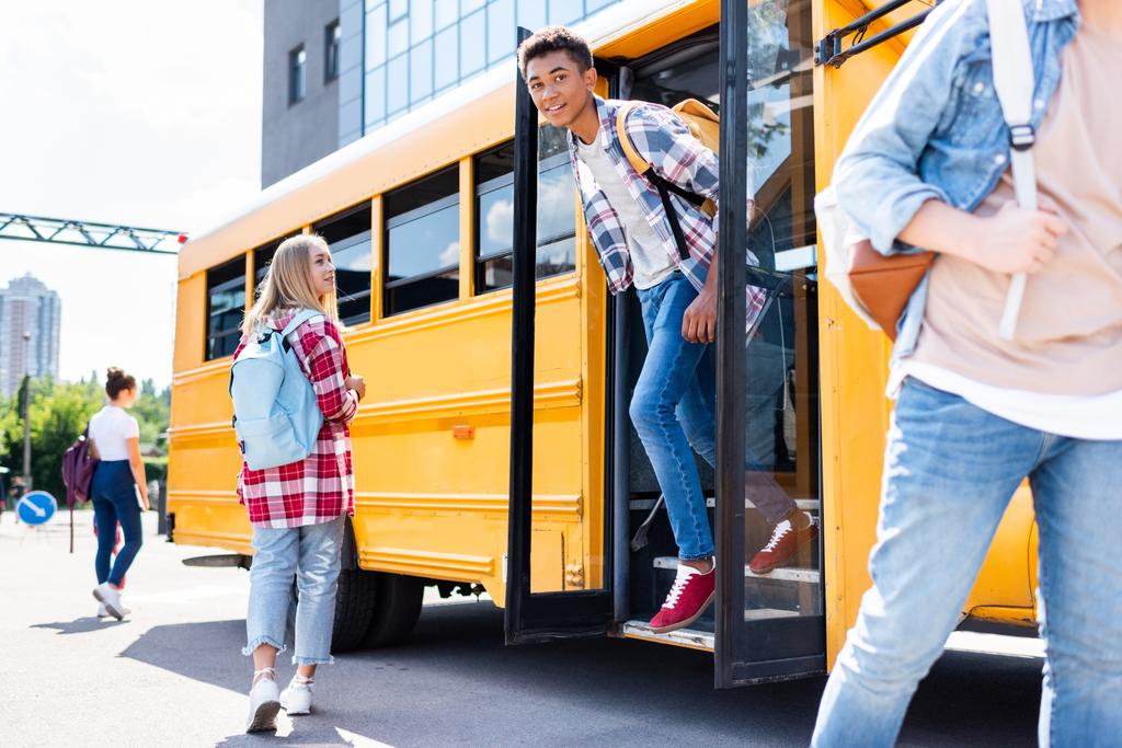 sonriente afroamericano adolescente escolar caminando fuera de la escuela autobús ingenio compañeros de clase alrededor
 - Foto, Imagen