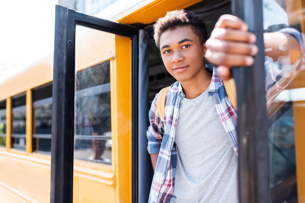 primer plano retrato de feliz adolescente afroamericano escolar de pie en la puerta del autobús escolar y mirando a la cámara
 - Foto, Imagen