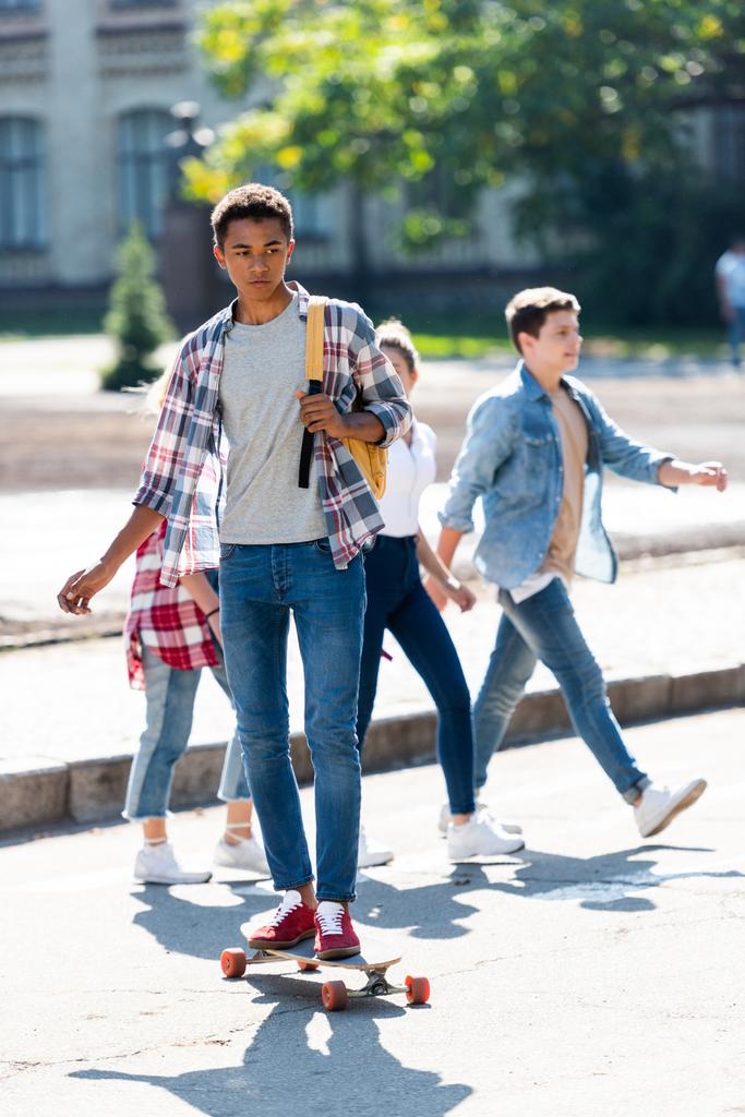 beau jeune écolier afro-américain chevauchant skateboard avec des camarades de classe flous marchant sur fond
 - Photo, image
