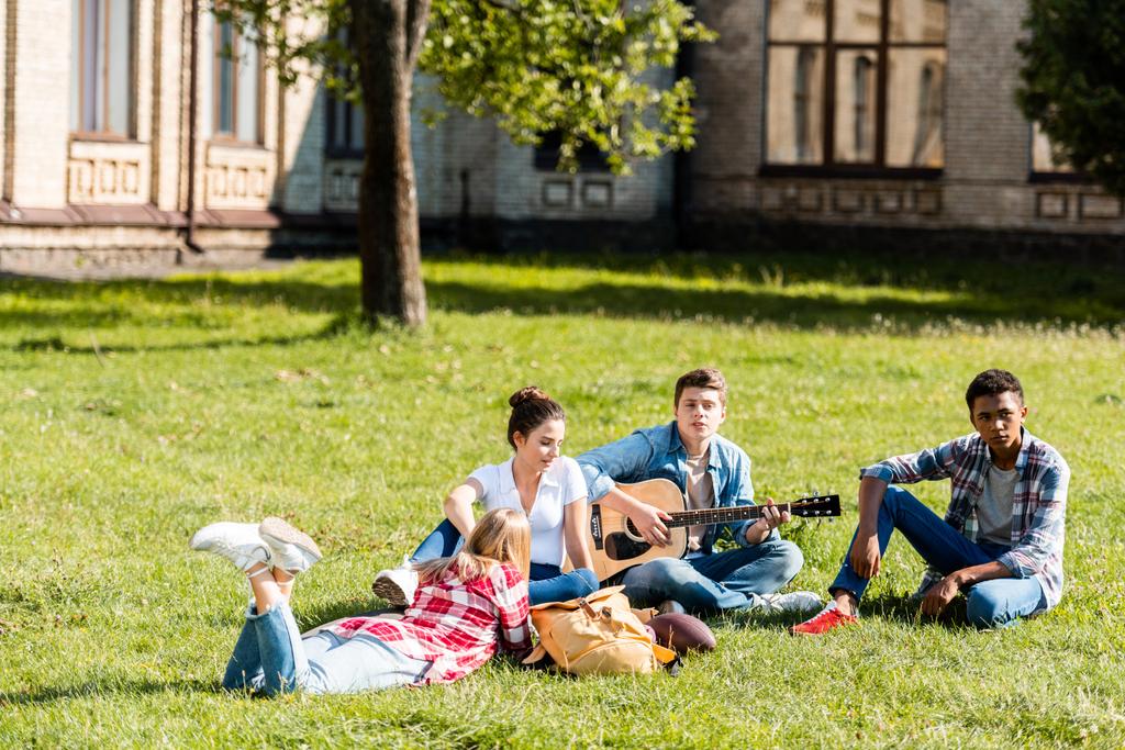 groep tieners samen tijd doorbrengen en luisteren naar gitaar liedje op gras - Foto, afbeelding