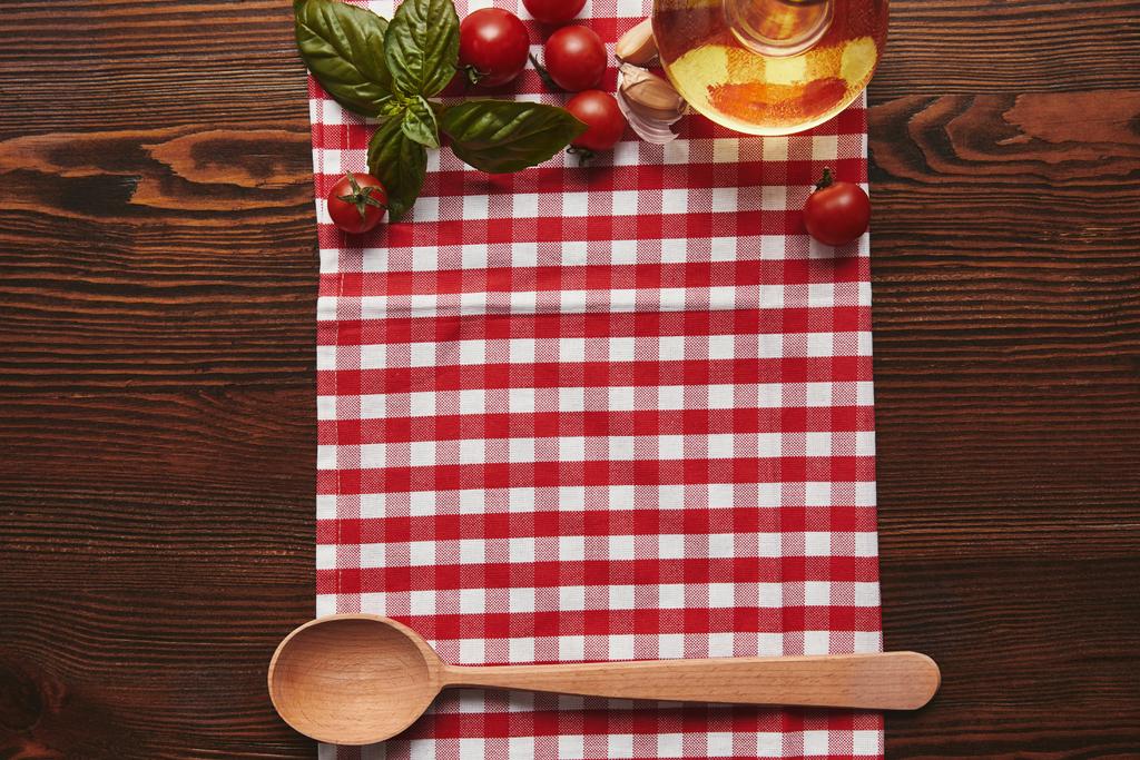 top view of checkered tablecloth, wooden spoon and fresh basil with tomatoes and oil on wooden surface - Photo, Image