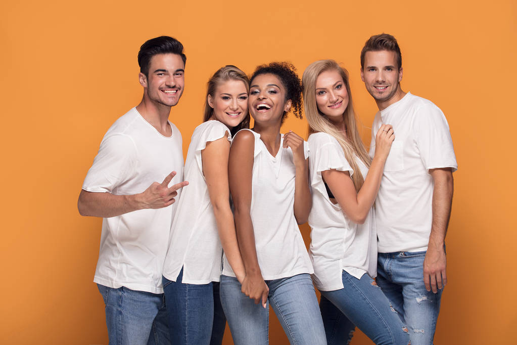 Vijf jonge multi etnische mensen glimlachen naar de camera, die zich voordeed op oranje studio achtergrond in witte casual shirts. - Foto, afbeelding