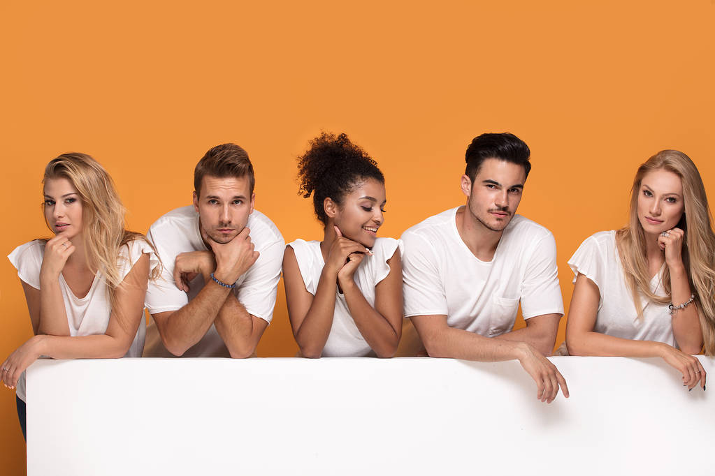 Группа молодых многонациональных красивых людей в белых рубашках, позирующих вместе с пустой белой доской. Веселые улыбающиеся друзья
. - Фото, изображение
