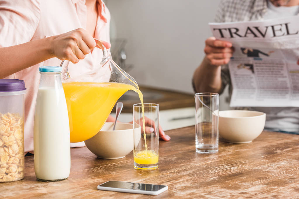 περικομμένη εικόνα κοπέλα ρίχνει χυμό σε γυαλί και ο φίλος που κατέχουν εφημερίδα στην κουζίνα - Φωτογραφία, εικόνα