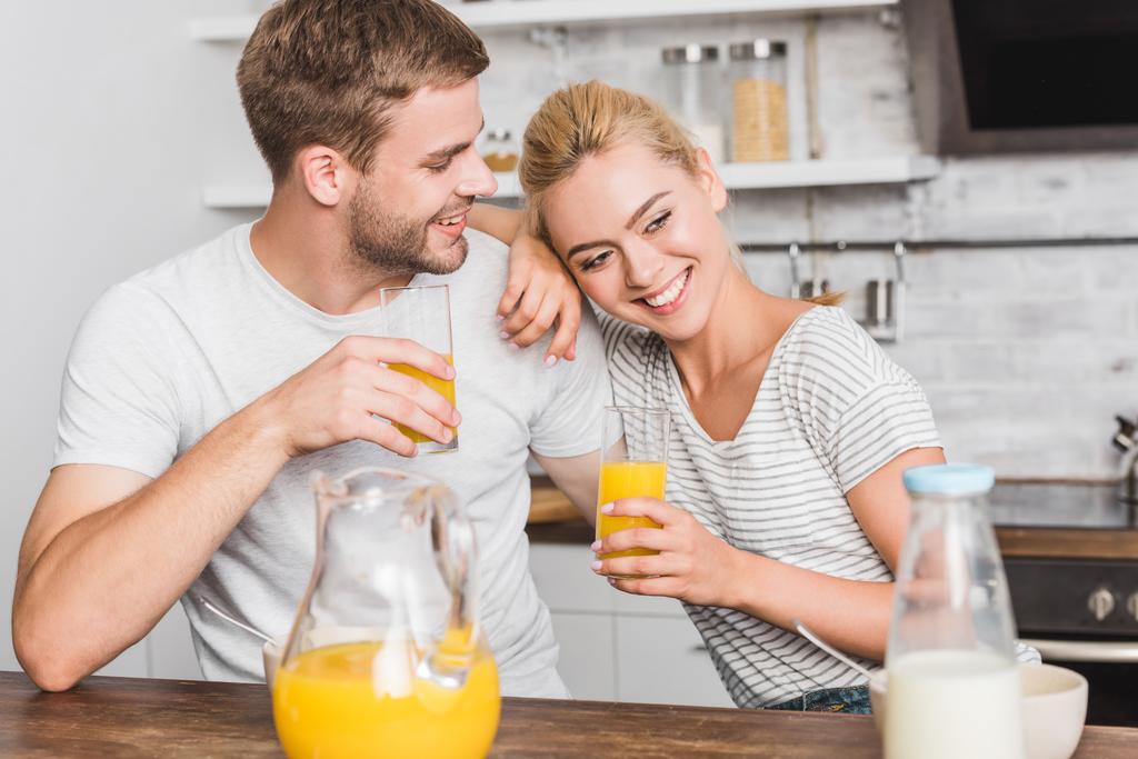 feliz pareja sosteniendo vasos de jugo de naranja y abrazos en la cocina
  - Foto, Imagen