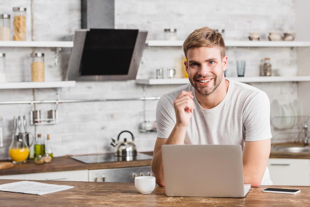 smiling handsome man working with laptop in kitchen and looking at camera - Photo, Image