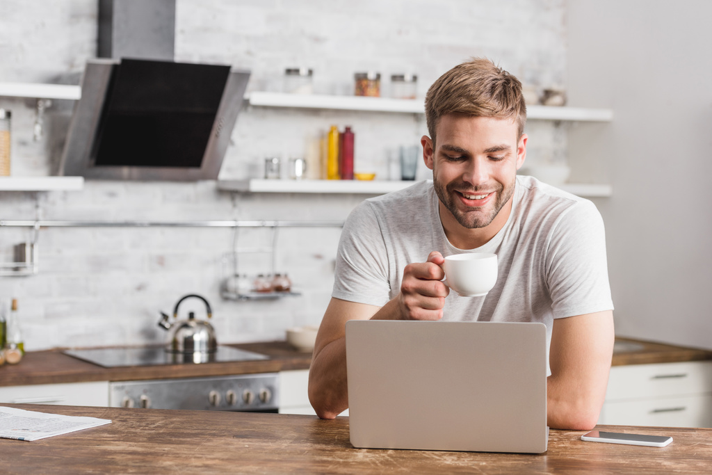 bonito sorrindo homem segurando xícara de café e olhando para laptop na cozinha
 - Foto, Imagem