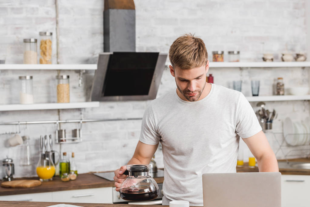 όμορφος άνδρας κρατώντας μπρίκι και κοιτάζοντας το laptop στην κουζίνα - Φωτογραφία, εικόνα