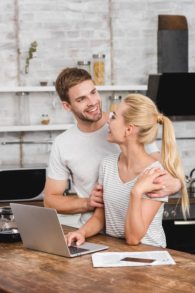 sorridente fidanzato abbracciare fidanzata in cucina mentre lei lavora con il computer portatile
 - Foto, immagini
