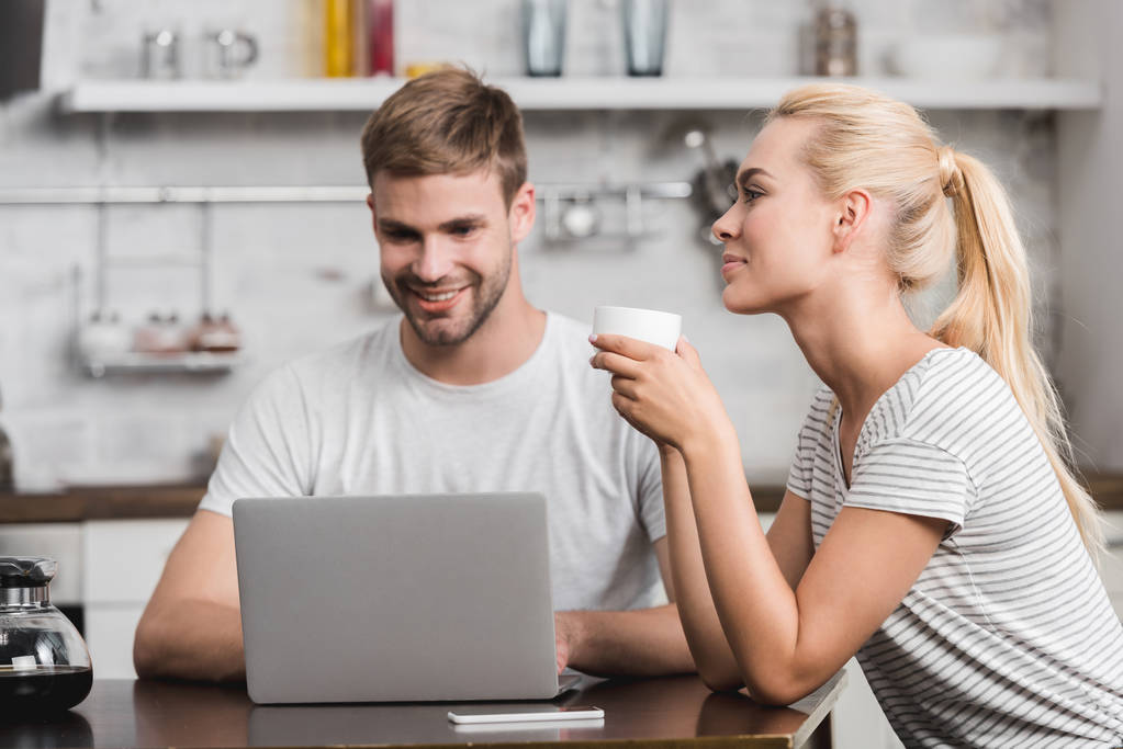 sonriente joven mujer sosteniendo taza de café mientras feliz novio usando portátil en casa
  - Foto, imagen