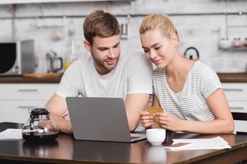 uśmiechnięta Młoda para trzymając wizytówkę i komputera przenośnego wspólnie przy stole w kuchni - Zdjęcie, obraz