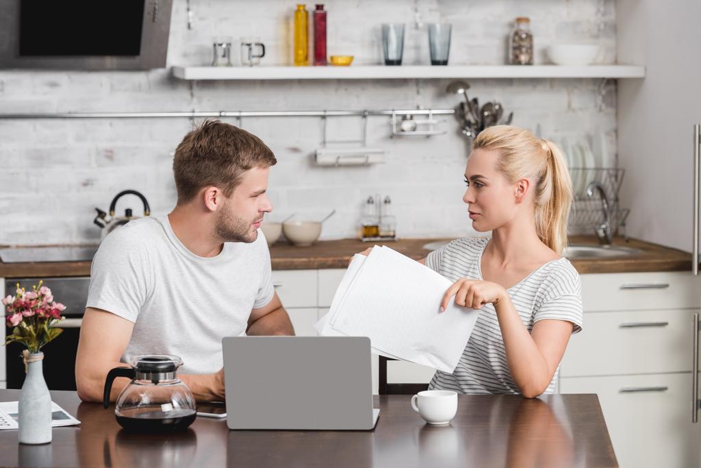 young couple holding papers and using laptop while sitting at kitchen table and looking at each other - Photo, Image