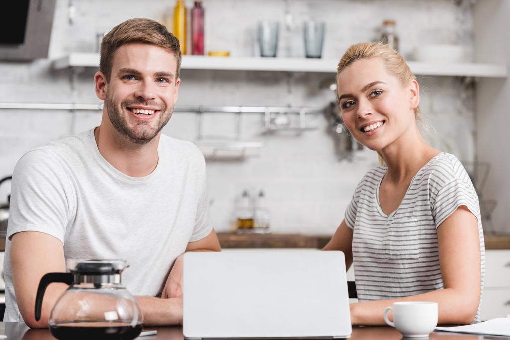 Happy νεαρό ζευγάρι χρησιμοποιώντας φορητό υπολογιστή και να χαμογελά στη φωτογραφική μηχανή μαζί  - Φωτογραφία, εικόνα