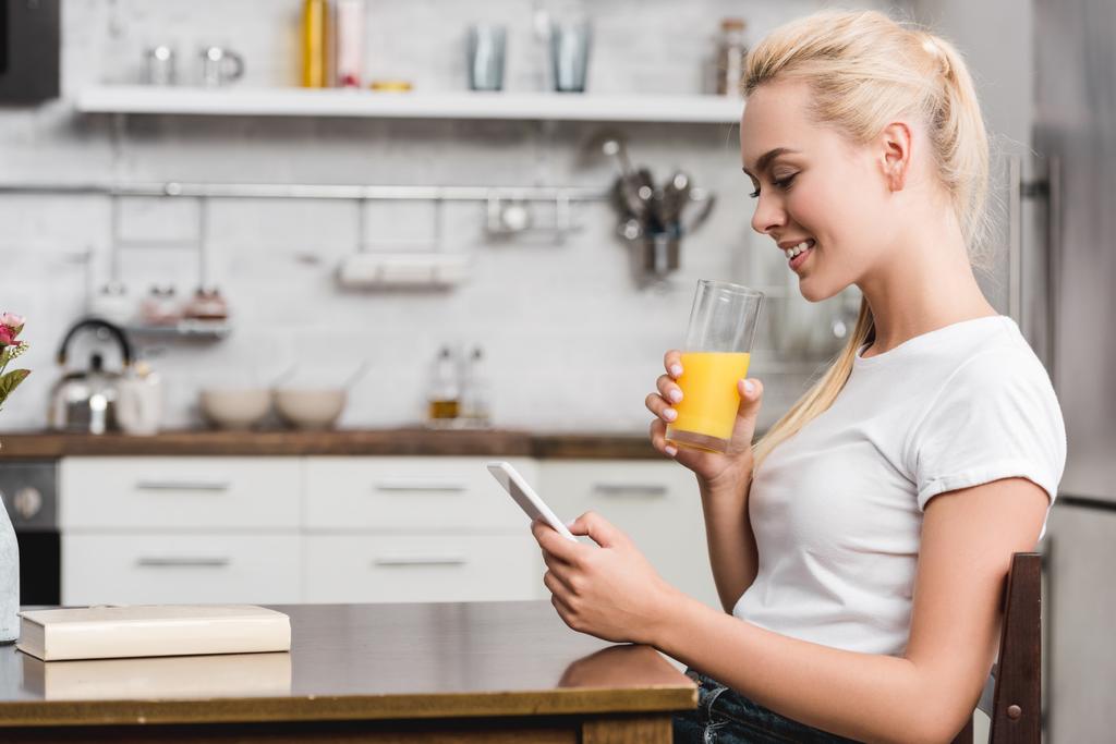 boční pohled na usměvavé mladé ženy pití čerstvé šťávy a použití smartphone doma - Fotografie, Obrázek