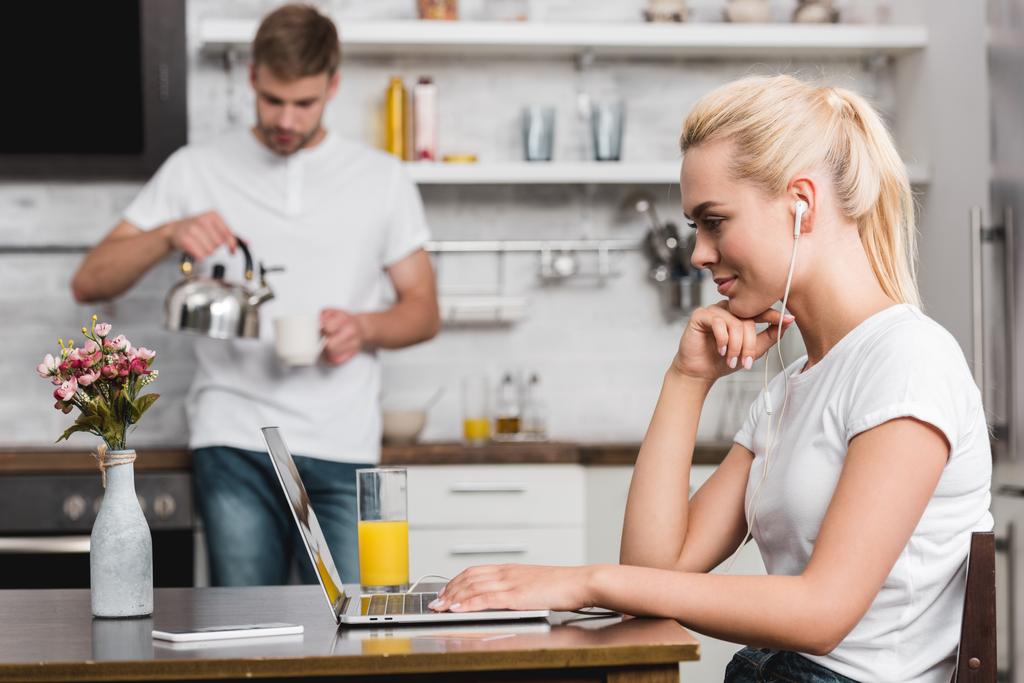 attractive young woman in earphones using laptop while boyfriend holding cup and kettle in kitchen  - Photo, Image