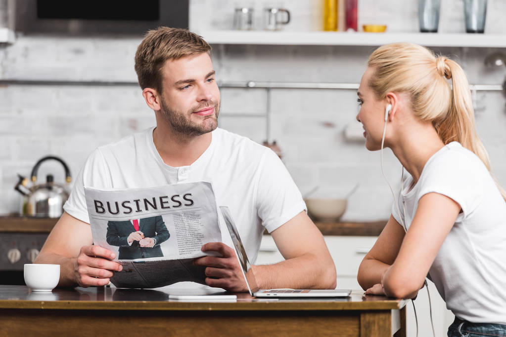 mosolygó fiatal férfi üzleti napilap olvasása és látszó-on boldog barátnője fülhallgató-konyha asztalnál ülve - Fotó, kép