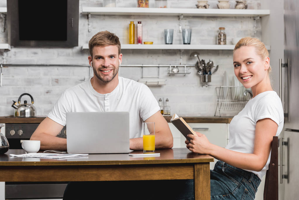 glückliches junges Paar mit Laptop und Buch am Küchentisch sitzend und lächelnd in die Kamera - Foto, Bild