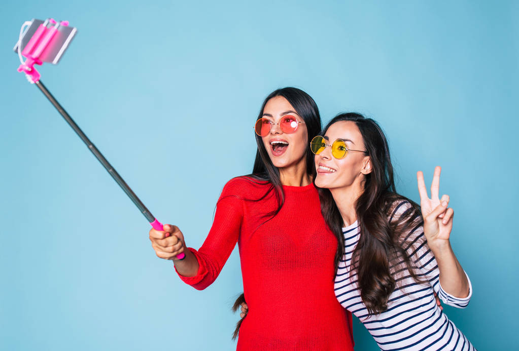 Portret van twee stijlvolle brunettes in zonnebril selfie met behulp van de selfie stick en smartphone op blauwe achtergrond maken - Foto, afbeelding