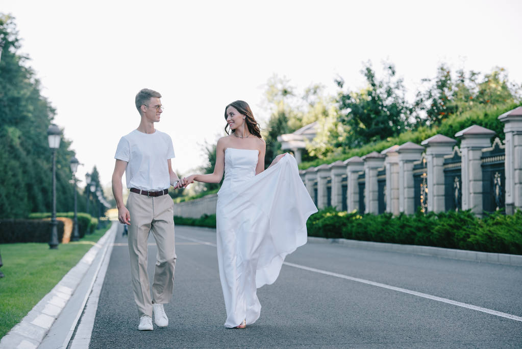 attractive bride and handsome groom holding hands and walking on road - Photo, Image