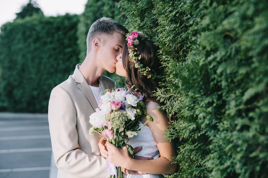 όμορφη γαμήλια ζευγάρι φιλιά και κρατώντας τη floral ανθοδέσμη  - Φωτογραφία, εικόνα