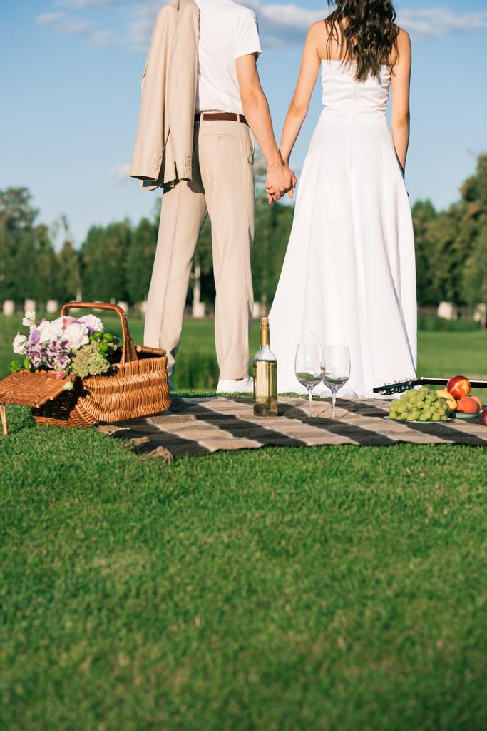 結婚式のカップルのピクニックの芝生の上に手を繋いでの背面図をトリミング - 写真・画像