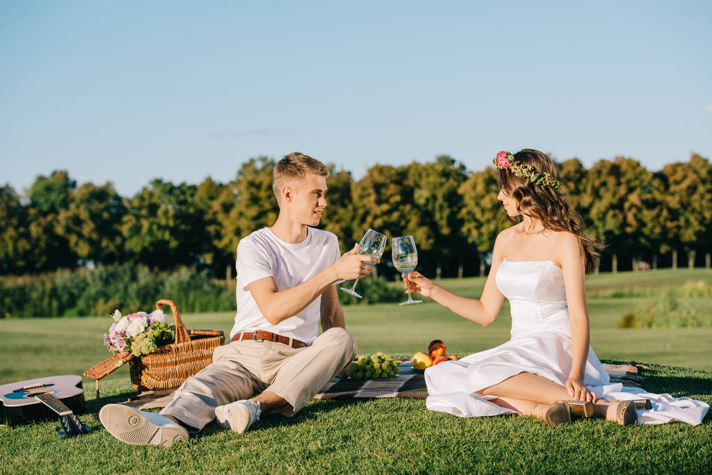 happy wedding couple clinking with glasses of wine during picnic on green lawn - Photo, Image