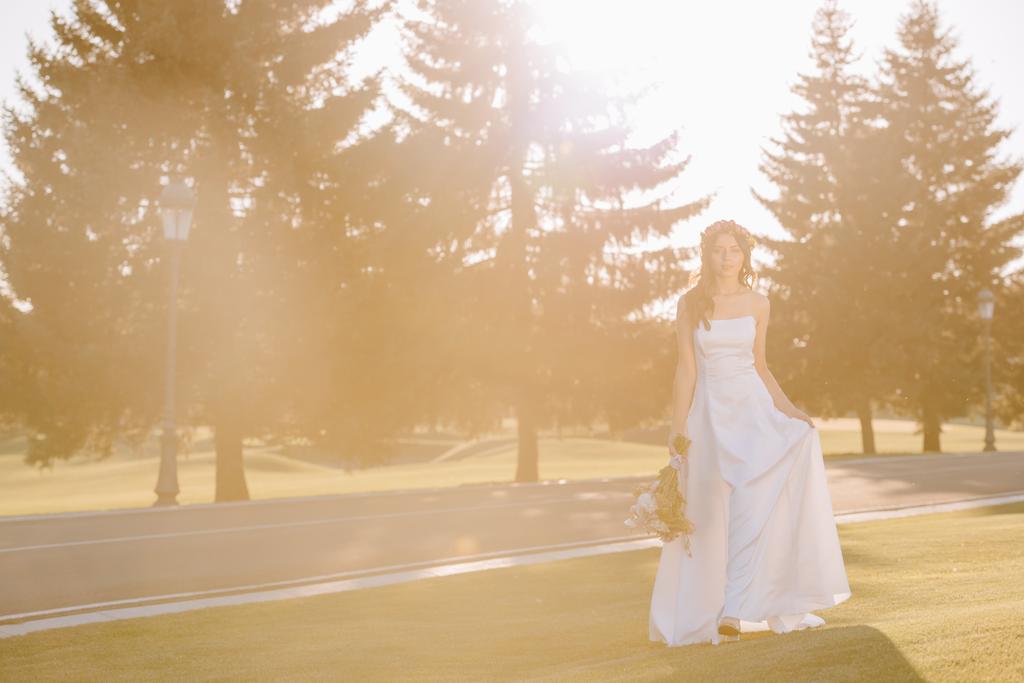όμορφη νύφη σε λευκό φόρεμα εκμετάλλευση γαμήλια ανθοδέσμη, σε εξωτερικούς χώρους με το πίσω φως - Φωτογραφία, εικόνα