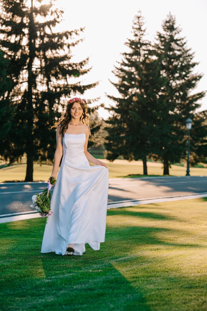 schöne glückliche junge Braut hält Hochzeitsstrauß und lächelt in die Kamera im Park - Foto, Bild